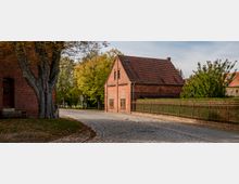 Gepflasterte Dorfstraße mit einem roten Backsteinhaus und einem großen Baum im Vordergrund. Im Hintergrund befinden sich gelb-grüne Laubbäume sowie ein Zaun und weitere Häuser.