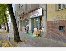 Straßenansicht mit einem kleinen Laden mit der Aufschrift „Beigeflüster“ auf einem weißen Schild. Vor dem Geschäft stehen Tische mit Tischdecken und Verkaufswaren, während links am Gehweg Bäume und Herbstlaub zu sehen sind.