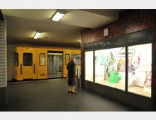 Unterirdische U-Bahn-Station mit einer gelben U-Bahn, die am Bahnsteig steht, und einem beleuchteten Schaufenster mit bunten Kleidungsstücken direkt neben einer älteren Frau, die es betrachtet. Die Wände sind mit Fliesen und dunklen Mosaikmustern verkleidet.