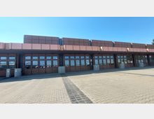 Flacher Gebäudekomplex mit brauner Fassade und großen, rechteckigen Glasfenstern. Der gepflasterte Vorplatz ist weitläufig, und der Himmel ist klar und blau.