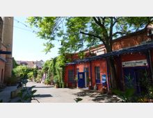 Innenhof mit roten Backsteingebäuden und blauen Türen und Fensterrahmen, teils mit Efeu bewachsen; an einer Fassade hängt ein Schild „Kurt Mühlenhaupt Museum“. Über dem gepflasterten Weg spannen sich Lichterketten, daneben stehen viele Pflanzkübel und ein großer Baum wirft Schatten.