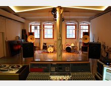 Tonstudio mit einem großen Mischpult im Vordergrund sowie mehreren Schlagzeugen, Lautsprechern und akustischen Elementen im Hintergrund. Der Raum ist durch hohe Fenster mit weiß verputzten Wänden und sichtbaren Deckenbalken geprägt.