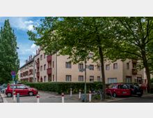 Wohnhaus in einer urbanen Umgebung mit hellbeigem Putz und roten Fensterrahmen sowie Balkonen. Im Vordergrund sind Autos, Fahrräder, Verkehrszeichen und Bäume zu sehen, die teilweise den Blick auf das Gebäude verdecken.