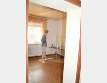 Ein einfacher Raum mit hellem Laminatboden, einer Holzkassettendecke und einer tapezierten Wand. Links befindet sich ein Fenster mit gemusterten Vorhängen und ein Heizkörper, daneben steht ein Tisch mit Geschirr und einem Wasserkocher vor einer gelb-weiß gefliesten Wand.