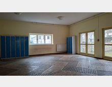Leerer Umkleide- oder Garderobenraum mit beigen Wänden, blau-grauen Schließfächern an den Seiten und einem Boden aus dunklen Ziegelplatten im Muster. An einer Wand befindet sich ein Fenster, daneben eine Glastür nach außen mit Blick auf eine Steinmauer.