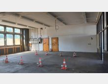 Leerer Innenraum einer Halle mit großen Fenstern auf der linken Seite und zwei orangefarbenen Türen an der weiß gestrichenen Rückwand. Im Raum stehen mehrere rot-weiße Absperrkegel auf dem Betonboden, Deckenlampen und Kabel hängen von der Decke.