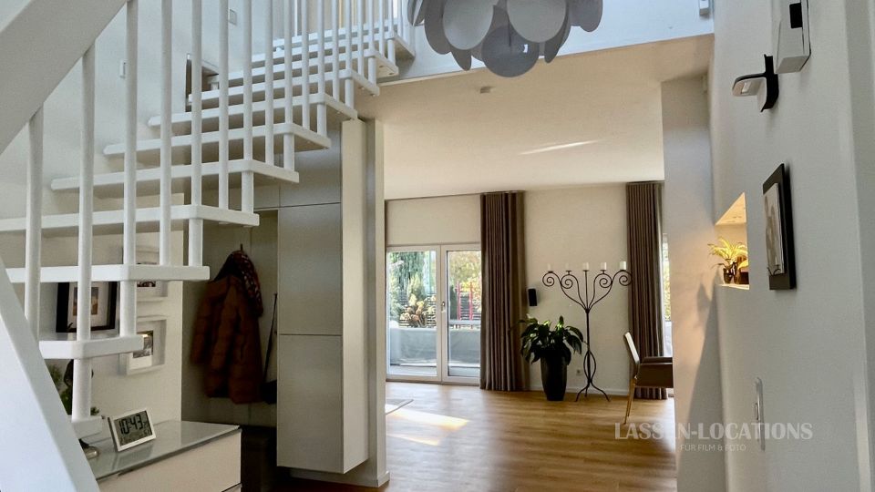 Heller, moderner Wohnhaus-Flur mit weißer Treppe und offener Galerie, hellem Holzboden und hoher Decke. Im Hintergrund führen große Terrassentüren mit Vorhängen zu einem Außenbereich; daneben stehen eine Zimmerpflanze und ein Garderobenständer.