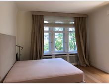 Ein helles Schlafzimmer mit einem großen Fenster, durch das Bäume sichtbar sind. Das Zimmer hat beige Vorhänge, helle Wände, einen Holzboden und ein Bett mit einem gepolsterten Kopfteil.