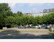 Ein kleiner Platz mit einer Baumschreihe im Vordergrund, Kiesboden und niedrigen Stufen. Im Hintergrund sind mehrstöckige Wohnhäuser und parkende Autos zu sehen.