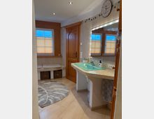 Badezimmer mit hellem Fliesenboden, einem türkisfarbenen Waschbecken auf einem gefliesten Waschtisch und einem großen Spiegel mit integrierter Beleuchtung. Im Hintergrund befinden sich eine Holztür, eine Sitzbank mit Kissen und geflochtenen Körben darunter sowie zwei Fenster mit halb geschlossenen Jalousien.