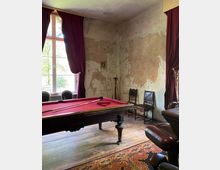 Ein rustikaler Raum mit einem Billardtisch mit rotem Tuch im Vordergrund, Holzboden und verputzten, abblätternden Wänden. Große Fenster mit roten Vorhängen lassen Tageslicht herein; im Hintergrund stehen Stühle und Billardzubehör an der Wand.