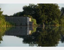 Holzhütte am Rand eines Sees, umgeben von dichtem Schilf und großen Bäumen, mit einem kleinen hölzernen Steg, der ins Wasser führt. Die ruhige Wasseroberfläche spiegelt die Umgebung deutlich wider.