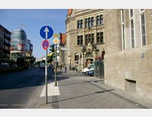 Straßenansicht mit einem historischen Gebäude rechts, verziert mit Ornamenten und großen Fenstern. Links befinden sich moderne Geschäftsgebäude, darunter ein rundes Einkaufszentrum mit der Aufschrift "Neukölln Arcaden". Straßenschilder und eine Ampel markieren die "Schönstedtstraße".