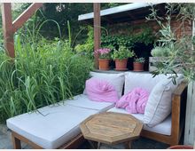 Gemütliche Sitzecke im Garten mit weiß gepolsterter Holz-Lounge und kleinem achteckigen Holztisch, umgeben von hohen Gräsern und Sträuchern. Im Hintergrund steht ein überdachter Holzschuppen mit Efeu und mehreren Blumentöpfen auf einem Regal.