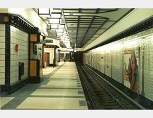 U-Bahn-Station mit weiß gefliesten Wänden, schwarzen Akzenten und rechteckigen Deckenleuchten. Ein Bahnsteig mit Gleis erstreckt sich in die Ferne, an den Wänden befinden sich Plakate und ein Schild mit der Aufschrift "Kiosk".