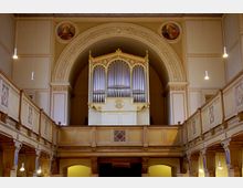 Innenansicht einer Kirche mit einer großen Pfeifenorgel auf einer Empore, die von reich verzierten architektonischen Elementen gerahmt ist. Die Wände und Decken sind mit Ornamenten und religiösen Gemälden geschmückt, und die Deckenleuchten hängen in regelmäßigen Abständen.