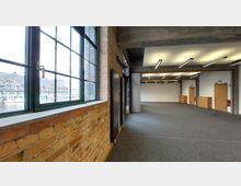 Großer Innenraum in einem modernisierten Industriegebäude mit freiliegenden Backsteinwänden, großen Fenstern mit Blick auf einen Kanal mit Gebäuden und Booten sowie sichtbaren Stahlträgern an der Decke. Der Raum ist mit grauem Teppich ausgelegt und verfügt über helle Wände, Holztüren und lineare Deckenleuchten.