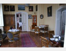 Historischer Salon mit mehreren Sitzgruppen und gedeckten Tischen, umgeben von antiken Möbeln und Porträts an den gelblich tapezierten Wänden; im Hintergrund führt eine geöffnete Doppeltür in einen angrenzenden Speiseraum mit eingedeckter Tafel.