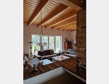 Wohnzimmer in einem Haus mit offenem Holzgebälk an der Decke, großen Fensterfronten, die auf eine Terrasse und in einen Garten blicken. Im Raum befinden sich ein Ecksofa, ein Couchtisch mit Tischdecke und Dekorationen sowie eine Stehlampe; rechts ist eine Steinwand sichtbar.