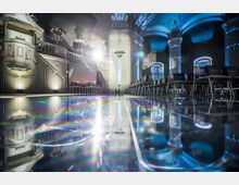 Innenraum einer historischen Halle mit gewölbter Decke und dekorativen Säulen, beleuchtet von blauen und weißen Scheinwerfern. Der glänzende Boden reflektiert die Umgebung, während Stuhlreihen an den Seiten aufgestellt sind; links steht eine verzierte Kanzel.