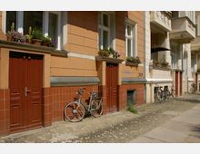 Fassadenansicht eines historischen Hauses mit ockerfarbenen Wänden und roten Holztüren, verziert mit Blumenkästen auf den Fensterbänken. Vor dem Gebäude stehen mehrere Fahrräder auf einem gepflasterten Gehweg.