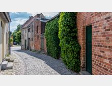 Schmale gepflasterte Gasse mit alten Ziegel- und Fachwerkhäusern, von denen einige mit Efeu bewachsen sind; im Hintergrund stehen Bäume und ein geparktes Auto.