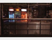 Eine Bar mit industriellem Design, bei der ein Gitter die Kühlschränke mit Getränken und bunte Beleuchtung im Hintergrund trennt. Im unteren Bereich ist eine perforierte Metallverkleidung sichtbar.