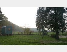 Ruhige Wiesenlandschaft mit Zaun und Bäumen, links ein kleiner grüner Bauwagen mit Fenster auf Holzstapeln. Im Vordergrund Rasen und ein gepflasterter Bereich, im Hintergrund Felder und eine bewaldete Hügelkette unter hellem Himmel.