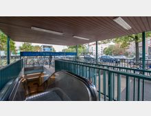 Eingang zur U-Bahn-Station "Amrumer Straße" mit Rolltreppen, umgeben von einem überdachten Bereich mit Holzdecke. Im Hintergrund sind ein Fahrradständer, geparkte Autos, Bäume und mehrstöckige Wohngebäude zu sehen.