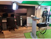 Tonstudio mit schalldämmenden Wänden, zwei großen Bildschirmen, einem Mischpult, schwarzen Bürostühlen und Beleuchtungseinrichtungen. Im Vordergrund ein Stehpult mit Laptop, Kabeln und Zubehör; im Hintergrund eine Puppe in Uniform und ein grüner Hintergrund.