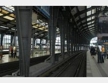 Bahnhofshalle mit überdachten Bahnsteigen, hohen stählernen Trägern und geschwungenem Glasdach; im Vordergrund Gleise und wartende Personen auf dem Bahnsteig, im Hintergrund beleuchtete Anzeigetafeln und eine Rolltreppe.