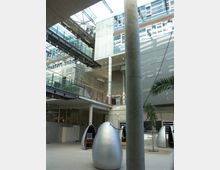 Modernes Gebäude mit einem offenen Atrium, durchzogen von Metallbrücken und großen Glasflächen. Im Erdgeschoss befinden sich eiförmige Metallkapseln, eine Palme und Sichtbeton-Elemente, die das industrielle Design unterstreichen.