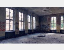 Verlassenes Gebäude mit großen, hohen Fenstern, die Licht in einen verwahrlosten Raum mit beschädigten Wänden bringen. Der Boden ist schmutzig, die Wände sind mit Graffiti bedeckt, und die Decke zeigt Spuren von Verfall.