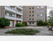 Wohnhof einer Plattenbausiedlung mit grauen Fassaden, Balkonen und Fenstern. Im Vordergrund ein gepflasterter Platz mit niedrigen Büschen, einem kleinen Sandkasten, einer Wippe und zwei Bänken, umgeben von Sträuchern und Bäumen.