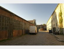 Industriegelände mit gepflastertem Hof, flankiert von Backsteingebäuden mit großen Fensterfronten auf der linken Seite und verputzten Gebäuden auf der rechten Seite. Im Hintergrund ist ein Torbogen sowie ein teilweise eingerüstetes Gebäude sichtbar, mehrere Fahrzeuge stehen im Hof.