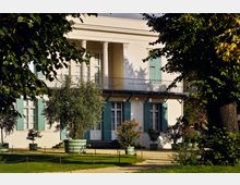 Klassizistische Villa mit heller Fassade, grünen Fensterläden und einem Balkon mit Säulen. Im Vordergrund ein Garten mit Rasenflächen, Bäumen und dekorativen Pflanzen in großen Blumentöpfen.