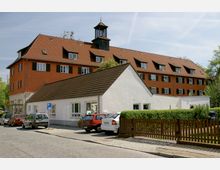 Mehrstöckiges Wohngebäude mit roten Dachziegeln, dunkelgrünen Fensterläden und einem kleinen Turm auf dem Dach im Hintergrund. Im Vordergrund ein einstöckiges weißes Gebäude mit Schrägdach, eine Reihe parkender Autos und ein Holzzaun, der eine Gartenfläche abgrenzt.