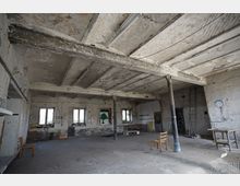 Großer, leerer Innenraum einer verlassenen Industriehalle mit roher Betondecke, freiliegenden Trägern und Stützsäulen sowie abblätternder Farbe an den Wänden. Mehrere Fenster lassen Tageslicht hinein; vereinzelt stehen einfache Stühle, ein Tisch und eine Leiter im Raum.