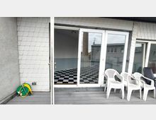 Eine Terrasse mit Holzboden und weißen Kunststoffstühlen, die an einer verglasten Tür grenzt. Durch die Tür ist ein Innenraum mit schwarz-weiß kariertem Boden und einer teilweise blau gestrichenen Wand sichtbar; an der Hauswand hängt ein aufgerollter Gartenschlauch.