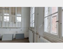 Ein leerer Raum mit weiß gestrichenen Wänden und großen, rechteckigen Sprossenfenstern, teils mit abblätternder Farbe. Im Raum befinden sich Heizkörper unter den Fenstern und graue Platten, die an der Wand lehnen.