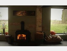 Ein rustikaler Innenraum mit einem schwarzen Kaminofen, in dem Feuer brennt, umgeben von einer grünen Wand mit sichtbaren Ziegeln. Auf beiden Seiten befinden sich große Fenster mit Blick ins Freie, Holzscheite in einem Korb und herbstliche Dekoration wie Kürbisse und Blumen.