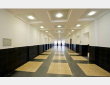 Langer, heller Flur in einem modernen Gebäude mit cremefarbenen Wänden und schwarzen Fliesen an den unteren Wandabschnitten. Der Boden besteht aus quadratischen, beige-schwarzen Fliesenmustern, und die Decke ist mit symmetrischen, eingelassenen Beleuchtungen versehen.