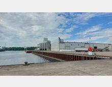 Industriegelände mit Lagerhallen am Rand eines Hafenbeckens. Im Vordergrund eine Betonpier mit rotbrauner Stahlkante am Wasser, im Hintergrund ein Bereich mit LKW, Lagergebäuden und Stapel von Säcken.