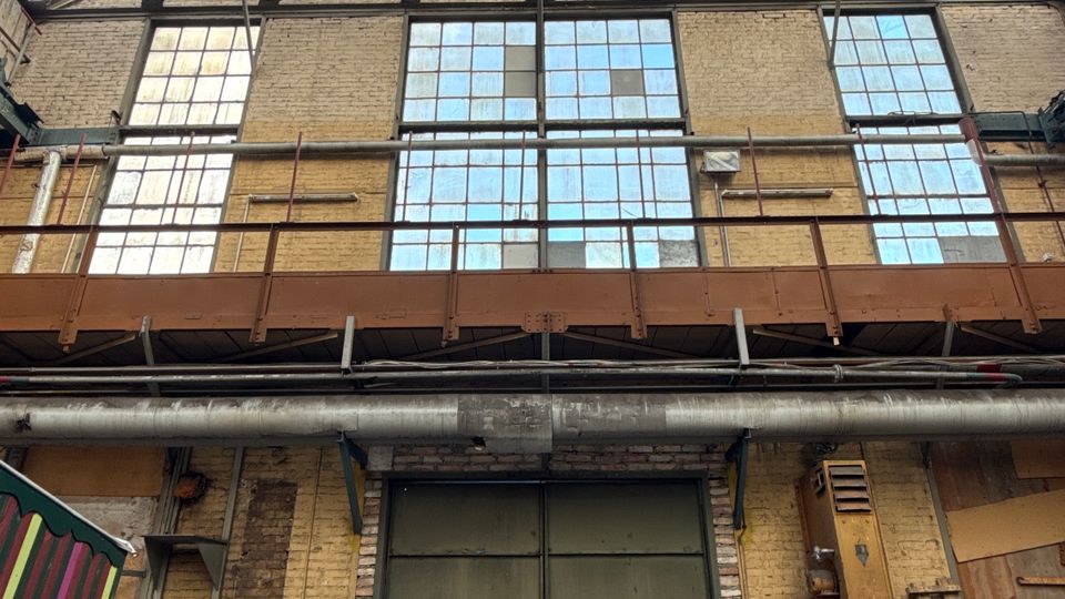 Innenansicht einer alten Industriehalle mit hohem Holzdach, Stahlträgern und großen Rasterfenstern entlang der Backsteinwand. Unten befindet sich ein großes, teilweise geöffnetes Metalltor mit sichtbaren Rohren und verstreuten Werkstattmaterialien am Rand.
