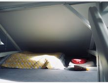 Eine niedrige Dachbodenfläche mit schräger Decke, einer Matratze mit gelb gemustertem Bezug auf dem Boden und einer Ansammlung von Yoga- oder Entspannungszubehör, darunter ein rotes Kissen und eine Schaumstoffrolle. Tageslicht fällt durch ein Fenster in die Szene.