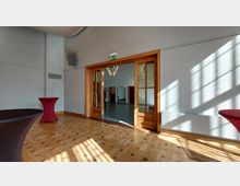 Heller Veranstaltungsraum mit Parkettboden, weißen Wänden und einer Glastür mit Holzrahmen, die in einen angrenzenden Raum führt. Im Vordergrund stehen zwei mit roten Stoffhussen bedeckte Stehtische, und an der Decke hängt ein Kronleuchter.