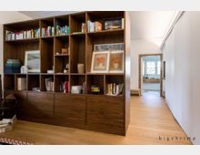 Ein Wohnraum mit einer großen Holzregalwand voller Bücher, gerahmter Bilder und Dekorationsobjekte. Im Hintergrund führt ein Flur mit Holzboden zu einem Badezimmer mit großem Fenster und weißen Fliesen.