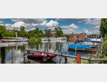 Bootsanlegestelle an einem ruhigen Fluss mit kleinen Motor- und Segelbooten, teils mit Planen bedeckt. Im Hintergrund grüne Bäume, ein historisches Backsteingebäude und weitere größere Boote; der Himmel ist leicht bewölkt.