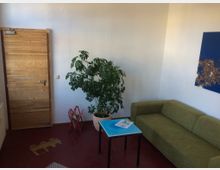 Ein kleiner Innenraum mit einer Holztür, einer grünen Zimmerpflanze in einem Topf, einem grünen Sofa und einem kleinen blauen Tisch. An der weißen Wand hängt ein abstraktes Kunstwerk mit blauem Hintergrund und goldfarbenen Details.