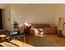 Wohnzimmer mit furniertem Holzboden und hellem beigen Teppich. Ein rosafarbener Samtsofa steht an der weißen Wand, daneben ein Couchtisch mit Büchern und eine hohe dekorative Lampe. Links daneben befindet sich ein schwarzer Stuhl aus Metall, ein Pflanzenständer mit einer Topfpflanze und eine geöffnete weiße Tür.